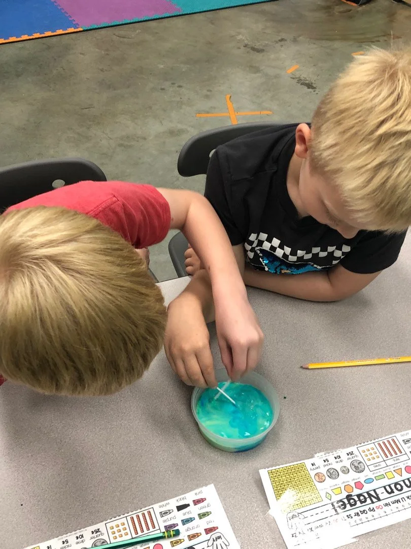 Two young boys with blonde hair are engaged in a science experiment at a table, mixing a blue and yellow liquid in a container, with a yellow pencil and educational sheets nearby.