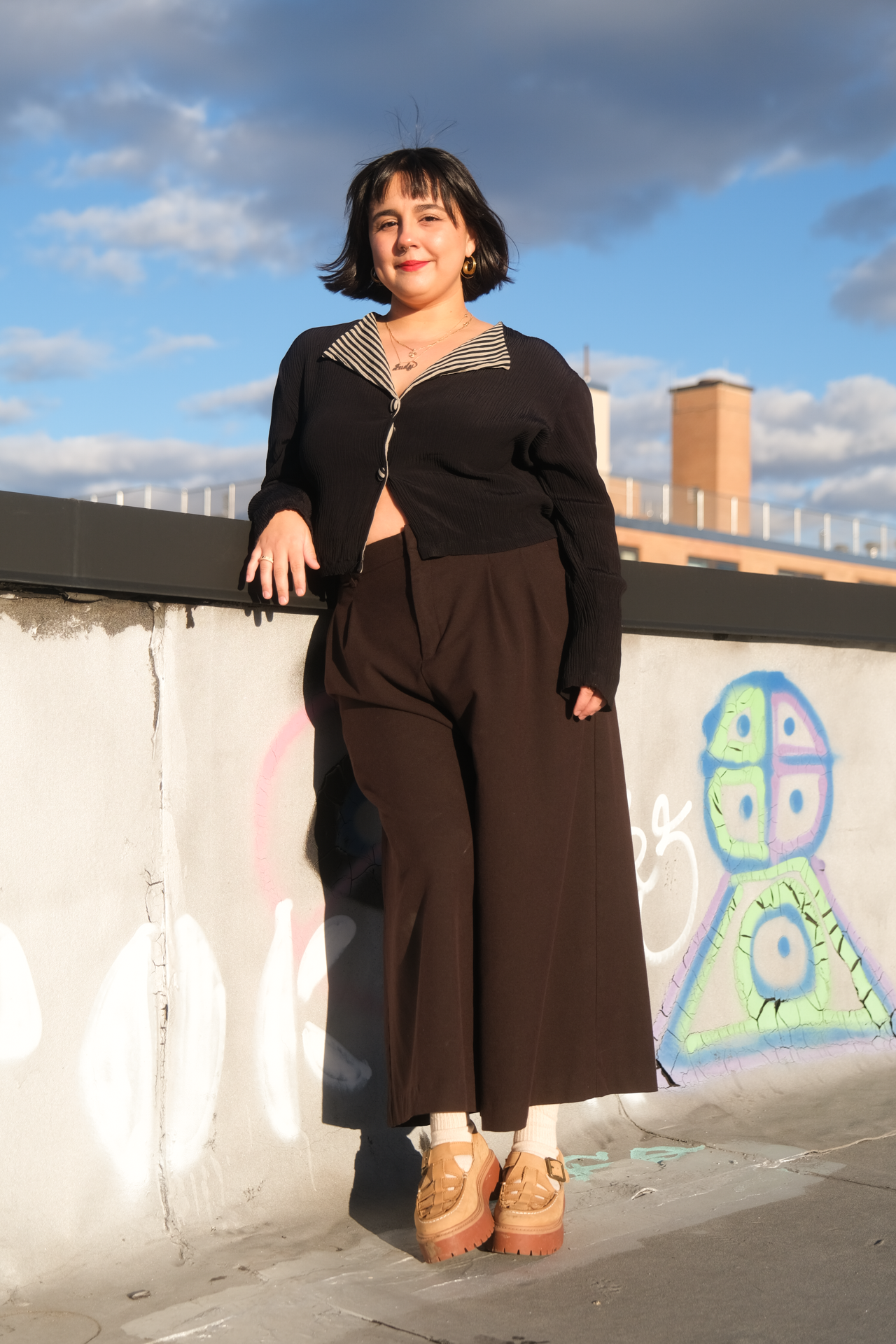 Aileen - short black hair in a black and beige striped top, wearing brown wide-leg pants and tan platform shoes, standing on a rooftop with graffiti art, under a partly cloudy sky.