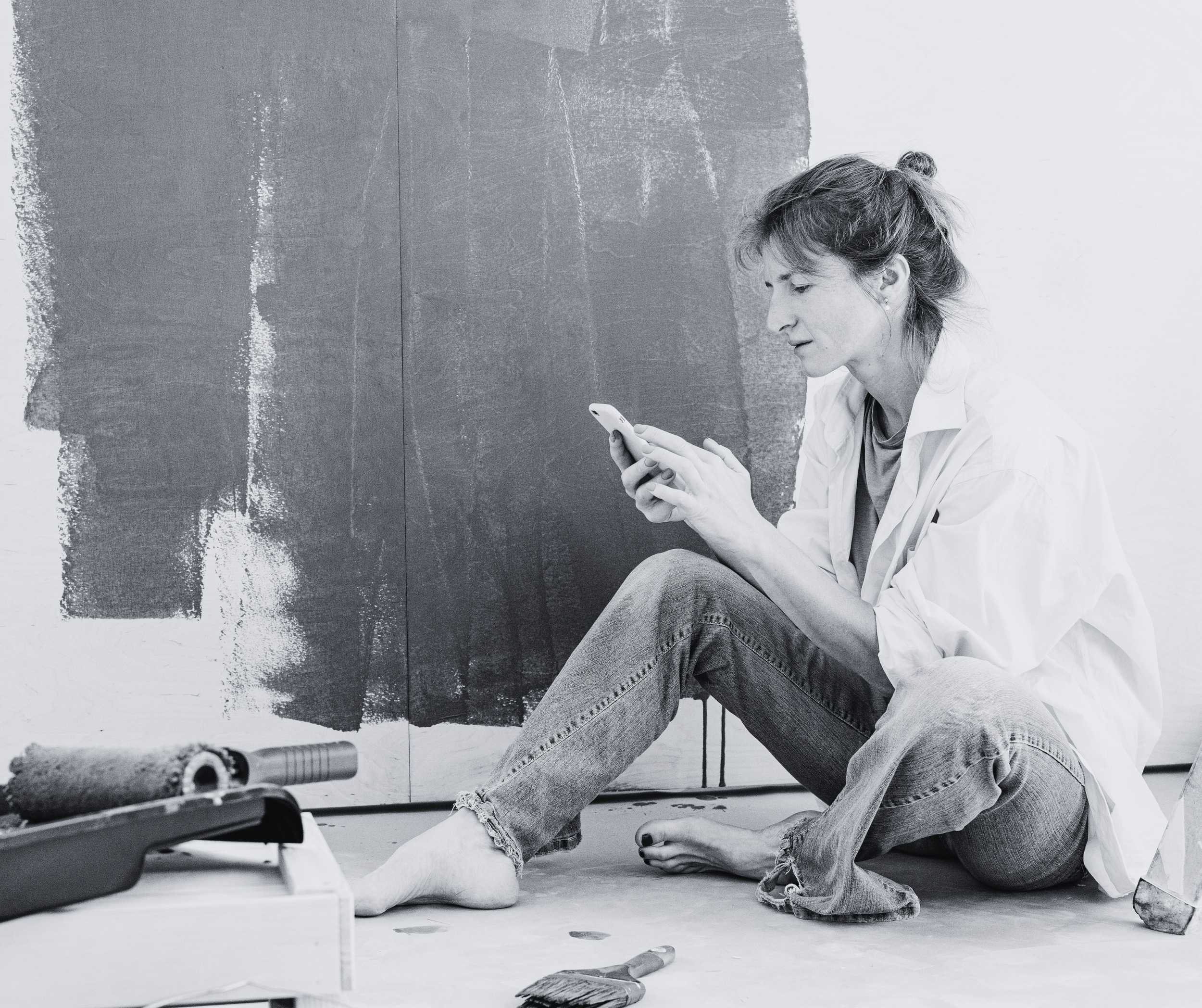 woman wearing jeans sits in front of a painting that is unfinishes while looking at her cell phone