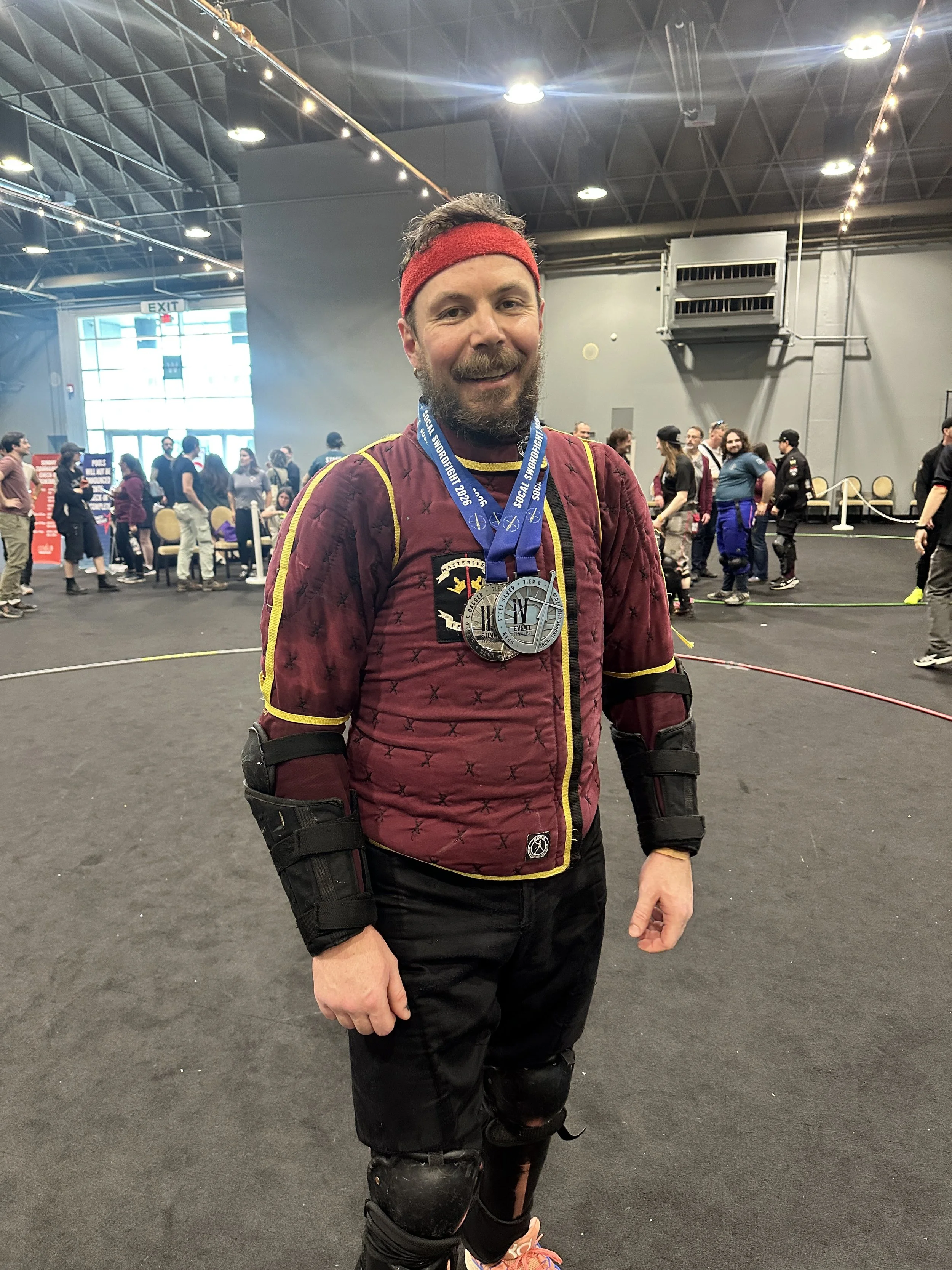 Man dressed in medieval-style fencing gear holding a sword inside a gym or dojo.