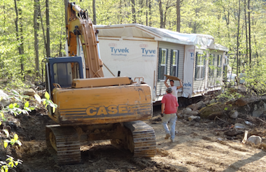The front piece squeezes through tight quarters as it fights its way through the soft ground of the driveway. The Case excavator pulls while the tow vehicle (mostly hidden) pushes. The combination has momentarily stopped while a worker places a mat ahead of the tires for them to roll over and help keep them from sinking into the ground.