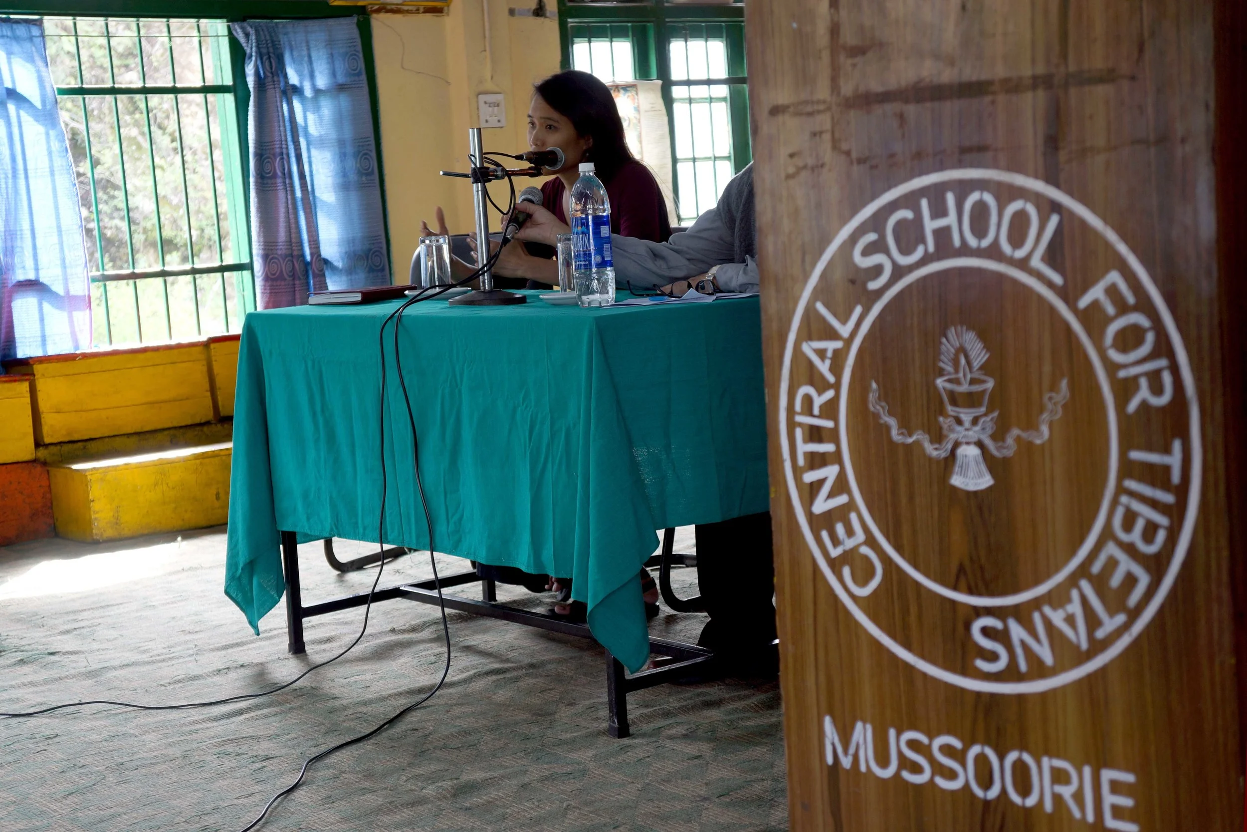 Two presenters give a talk. Next to them is a podium that reads "Central School For Tibetans, Mussoorie"