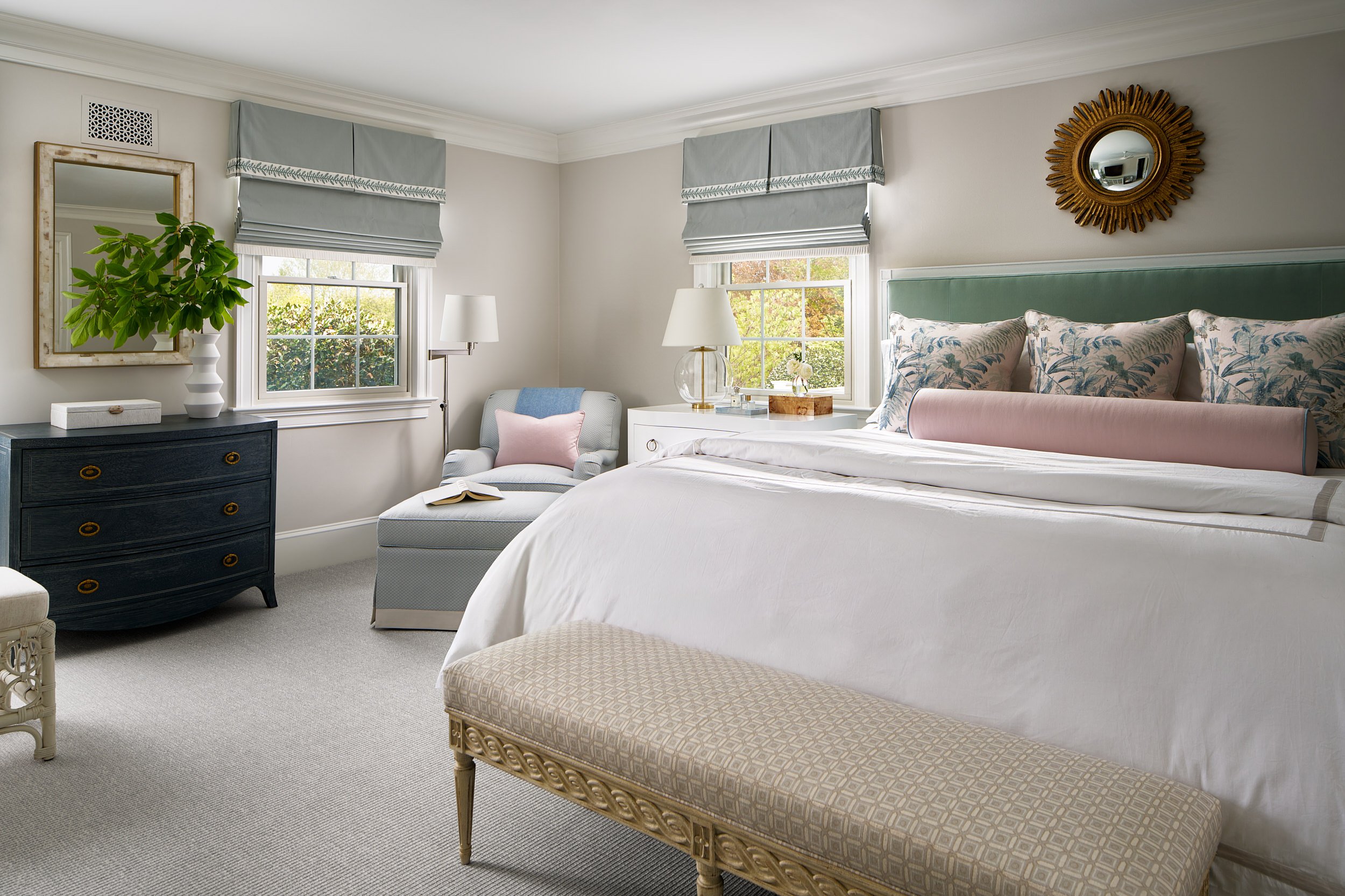Primary bedroom with a green upholstered headboard, pink bolster pillow, and a gold sunburst mirror, interior design photography in Maryland