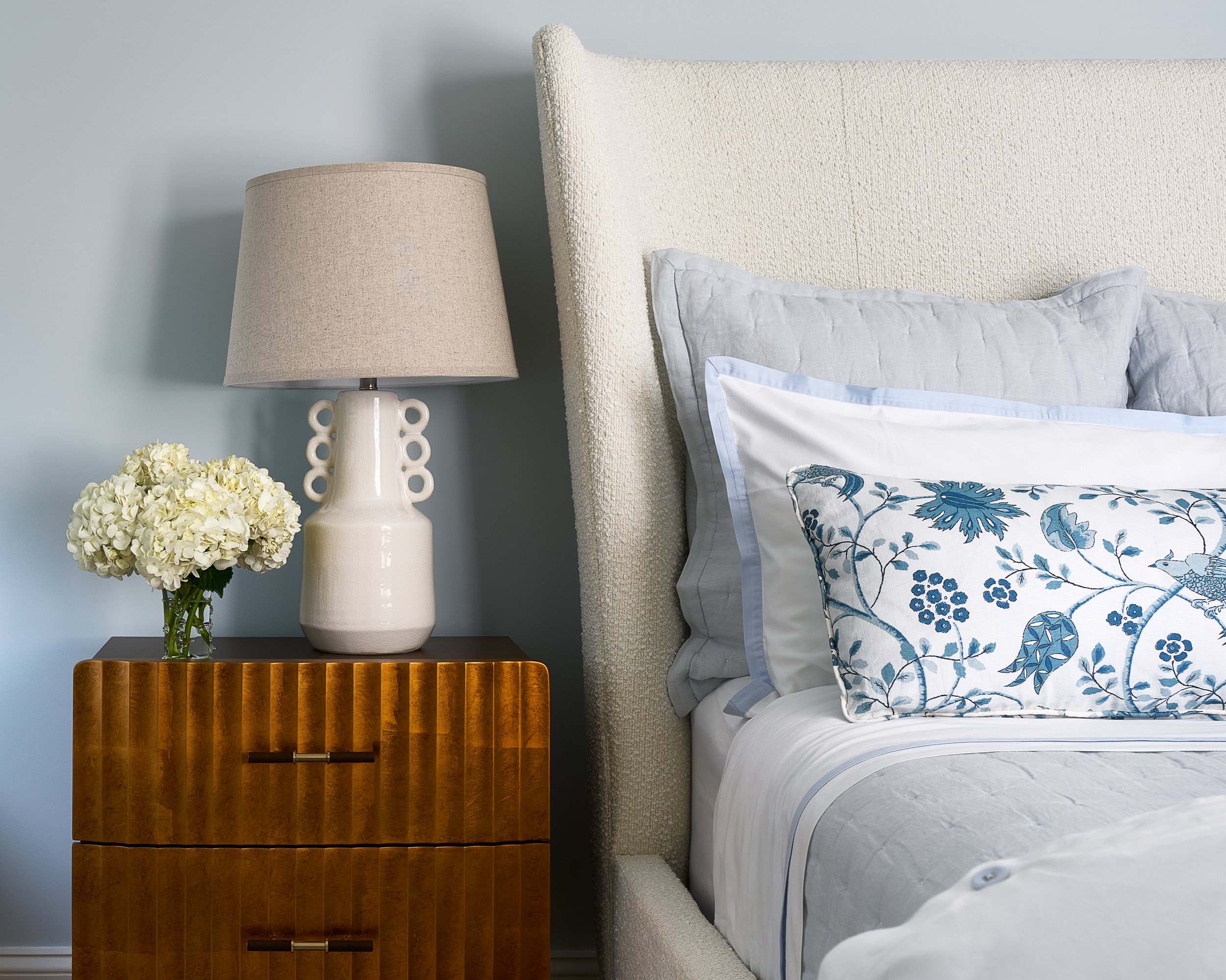 Bedroom vignette with a fluted wood nightstand, cream boucle headboard, and a blue floral accent pillow, interior design photography in the DMV area