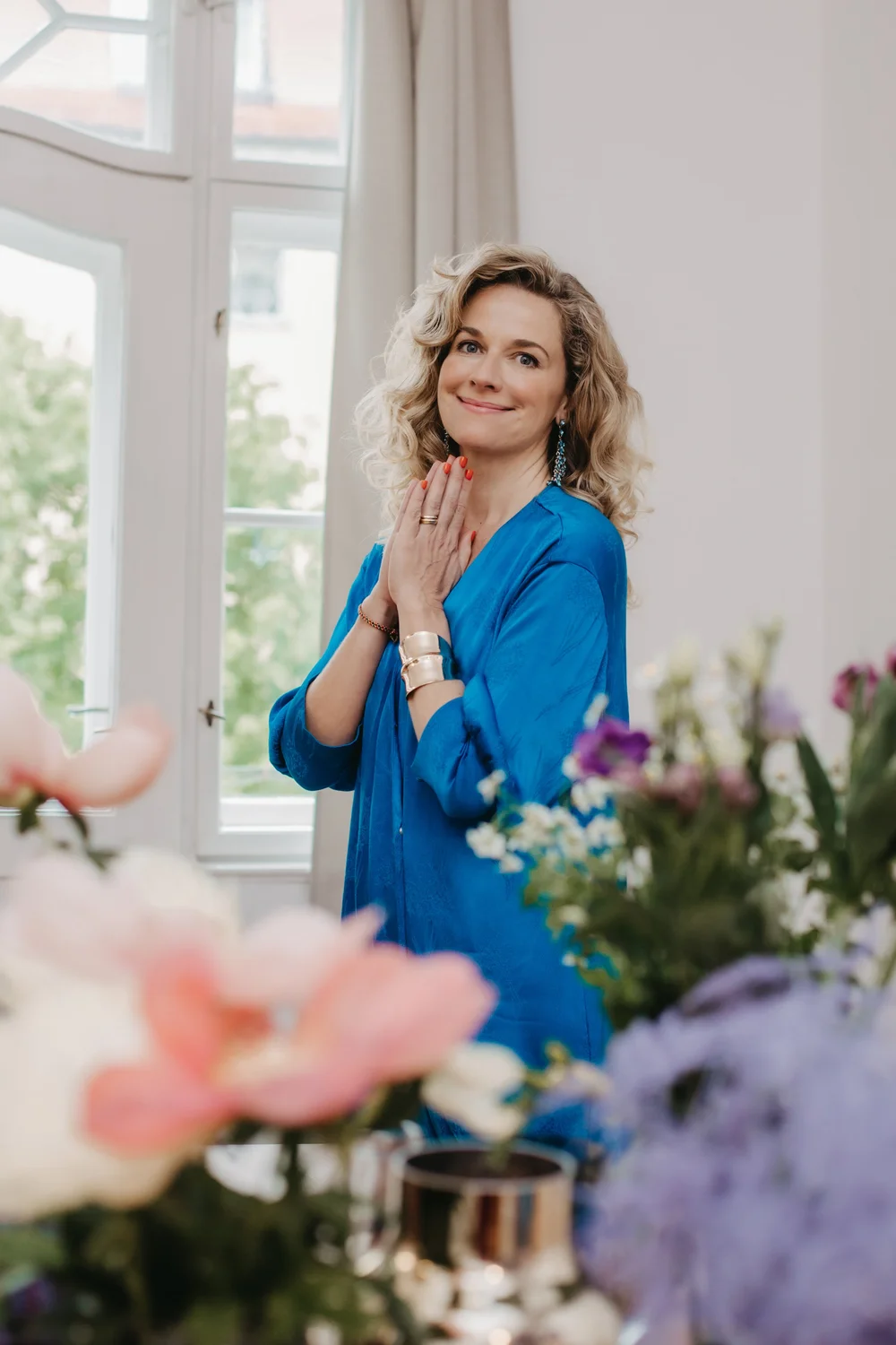 Kristina Sacken im blauen Kleid mit zusammengelegten Händen am Fenster