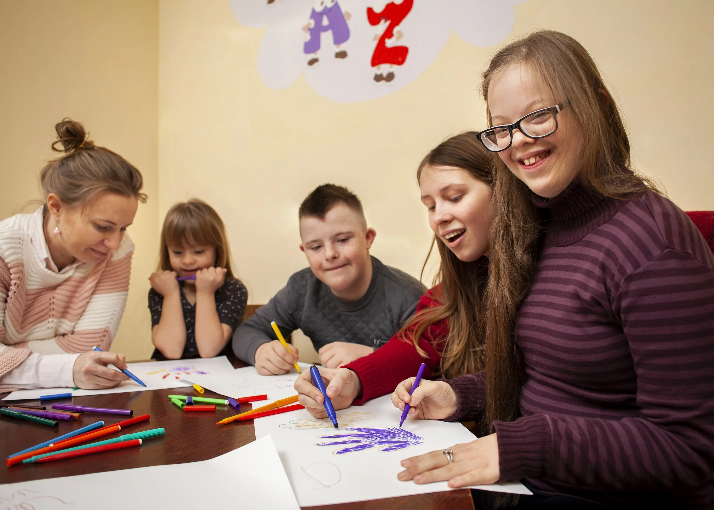 A group of smiling individuals, including young adults and a child, are gathered around a table drawing with colorful markers, conveying a joyful, inclusive atmosphere.
