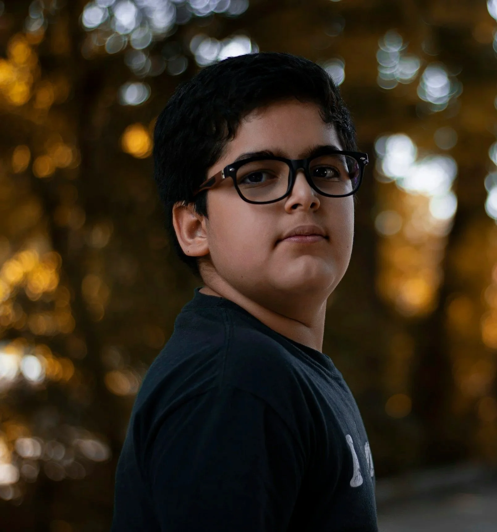 A boy wearing glasses gazes thoughtfully to the side, with a reflective double exposure effect. The background shows a warm, blurred tree canopy.