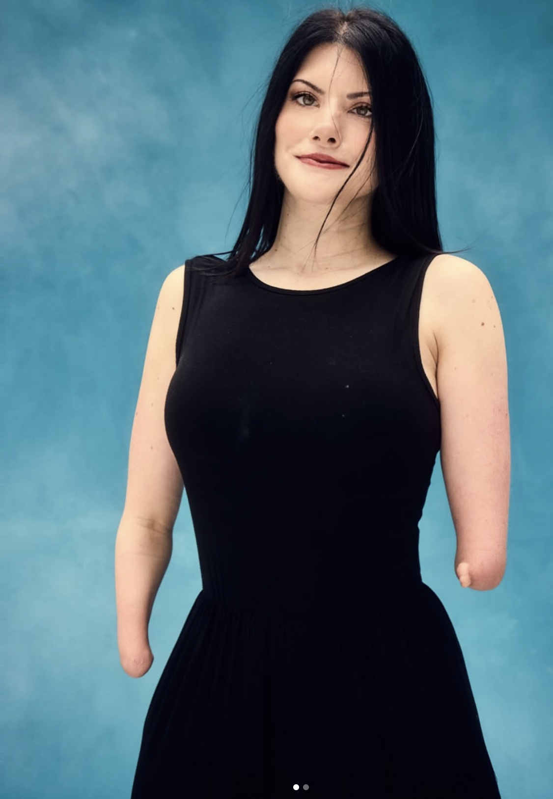 A person with long black hair stands in front of a blue studio backdrop, smiling slightly. They are wearing a sleeveless black dress and have bilateral upper-limb differences.
