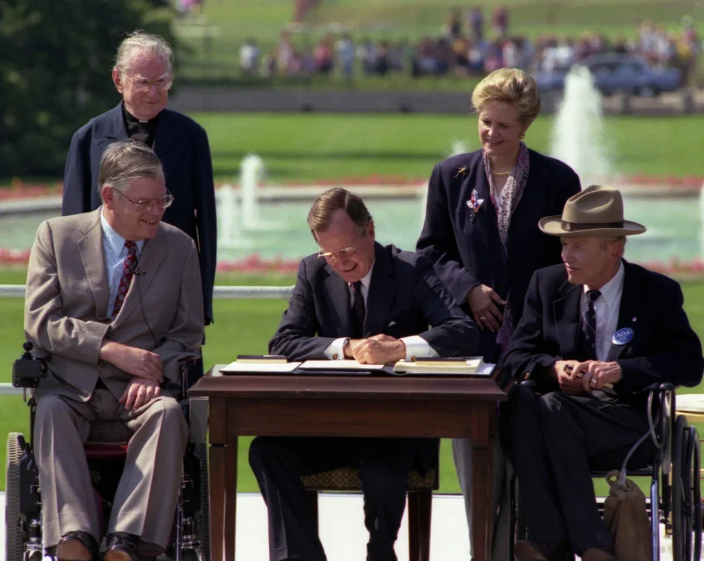 A group of five people gather outdoors at a signing ceremony. A man in a suit signs a document on a desk. Others watch, two sitting in wheelchairs.