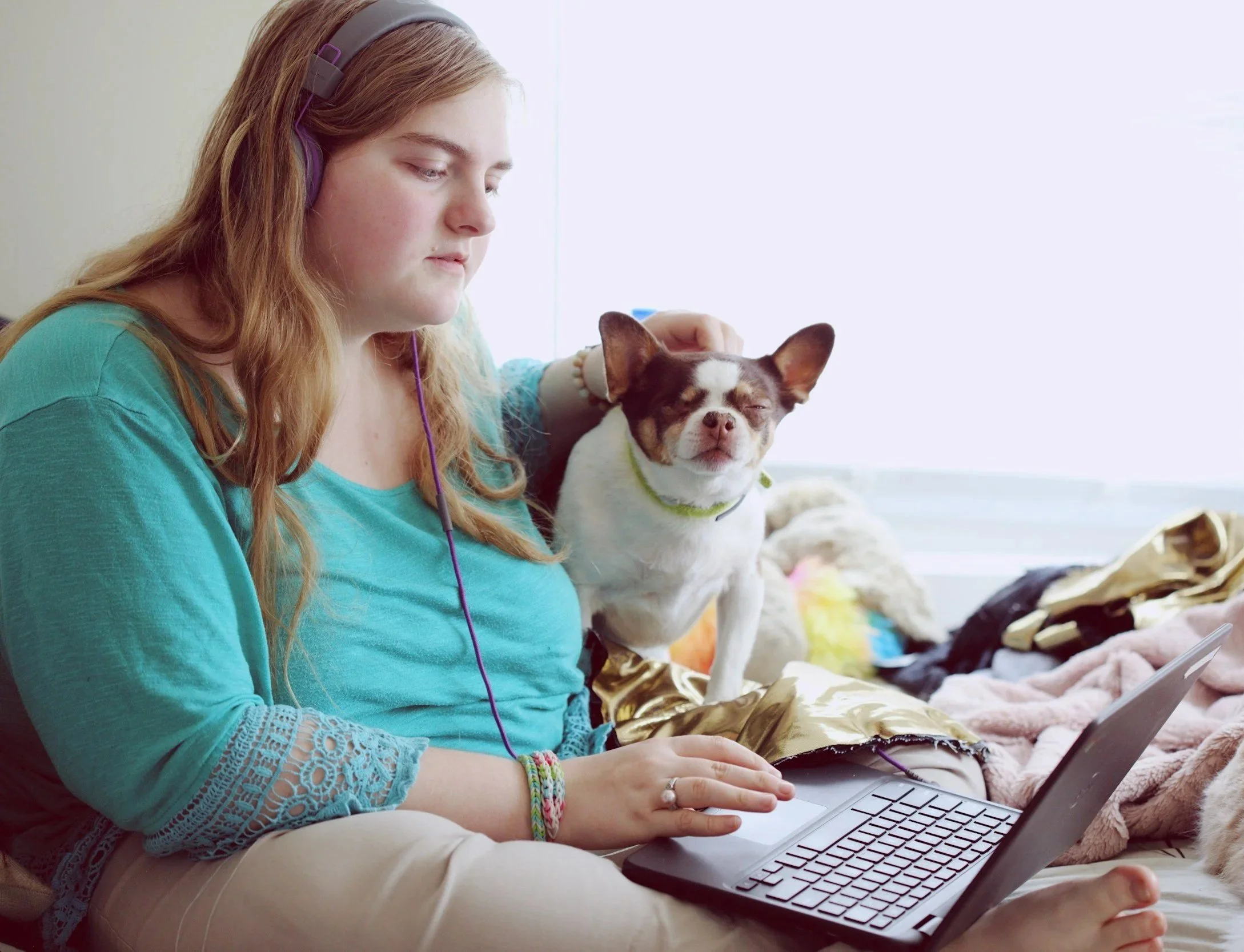 A woman wearing headphones sits near a window, focused on a laptop. She gently pets a small dog by her side, creating a calm, cozy atmosphere.