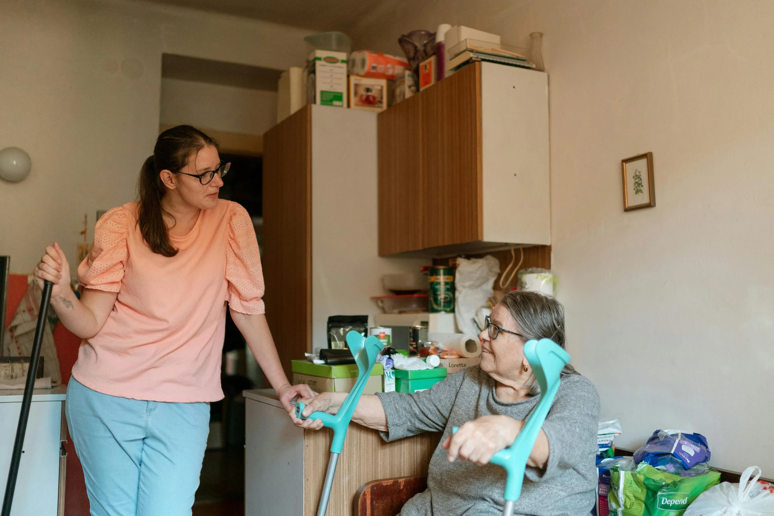 A young woman with glasses stands beside an elderly woman using crutches in a cozy room. They appear to be engaged in a warm conversation.