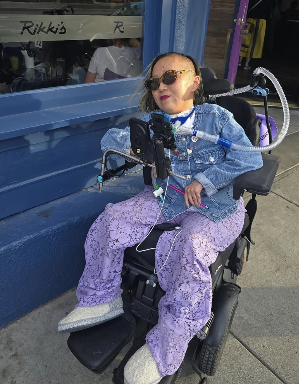 A smiling person in sunglasses sits in a powered wheelchair outside a storefront, wearing a denim jacket and lavender pants. Their wheelchair includes a mounted communication device, and they use a ventilator connected through a tracheostomy.