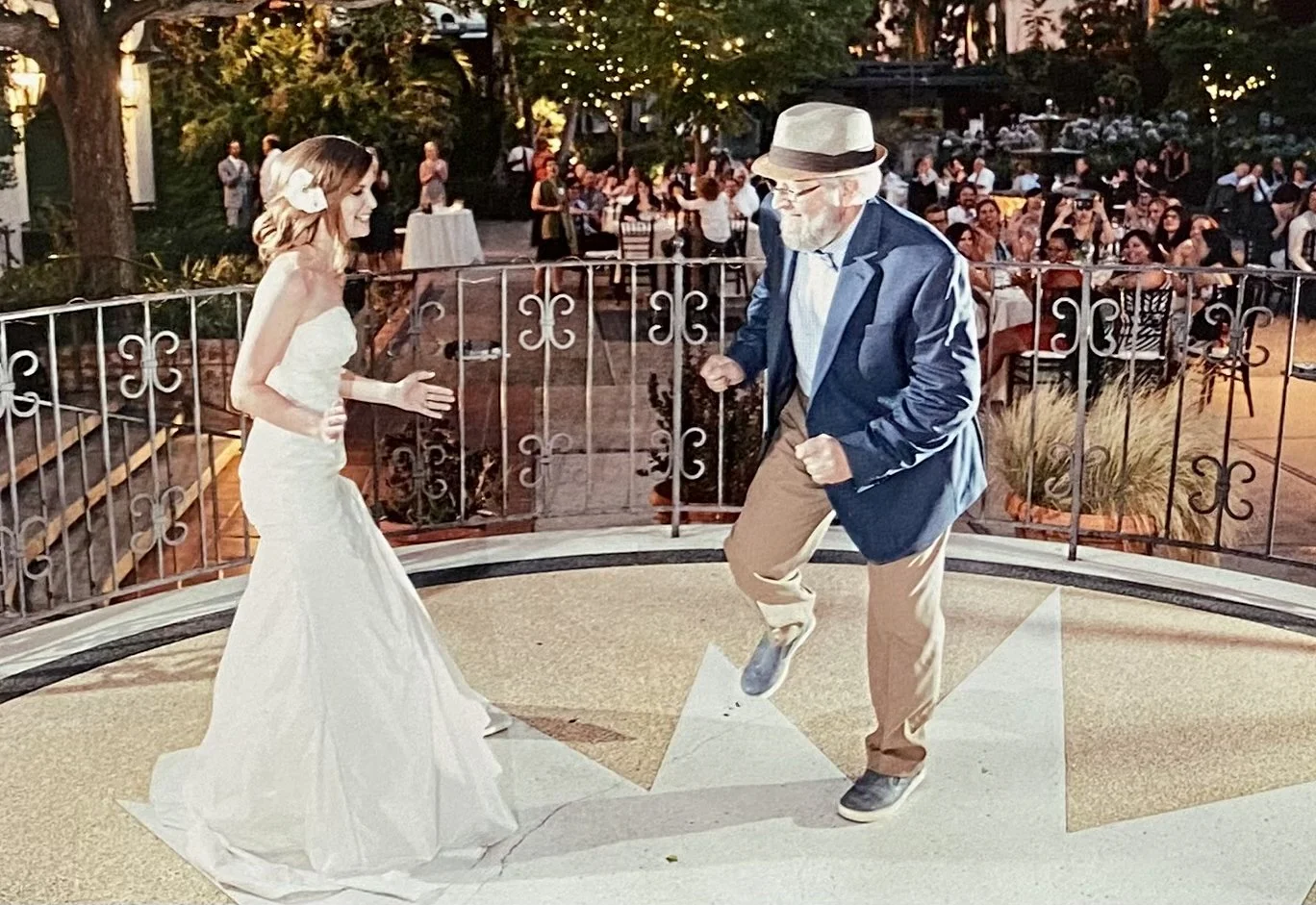 A woman in a white wedding dress dances with a man in a blue jacket and tan slacks on a outdoor raised platform in front of wedding guests.
