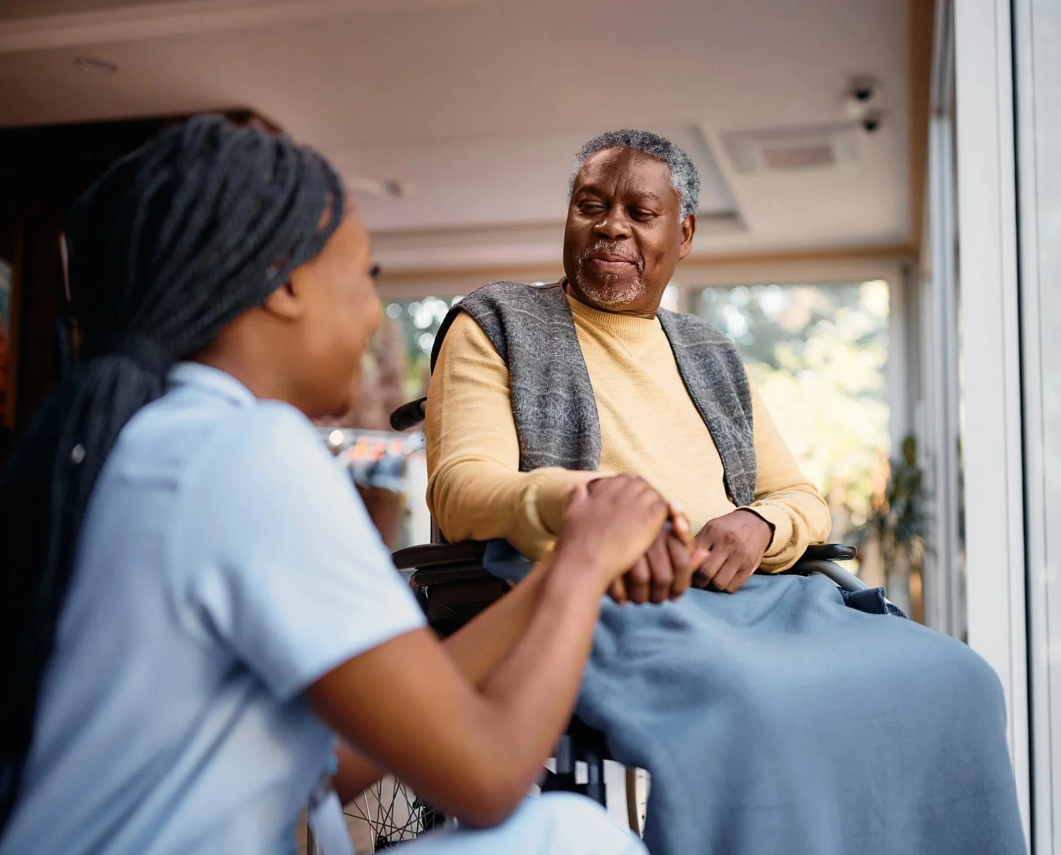 A caregiver kneels beside a wheelchair user indoors, sharing a warm, supportive moment. Sunlight filters through a window, creating a peaceful ambiance.