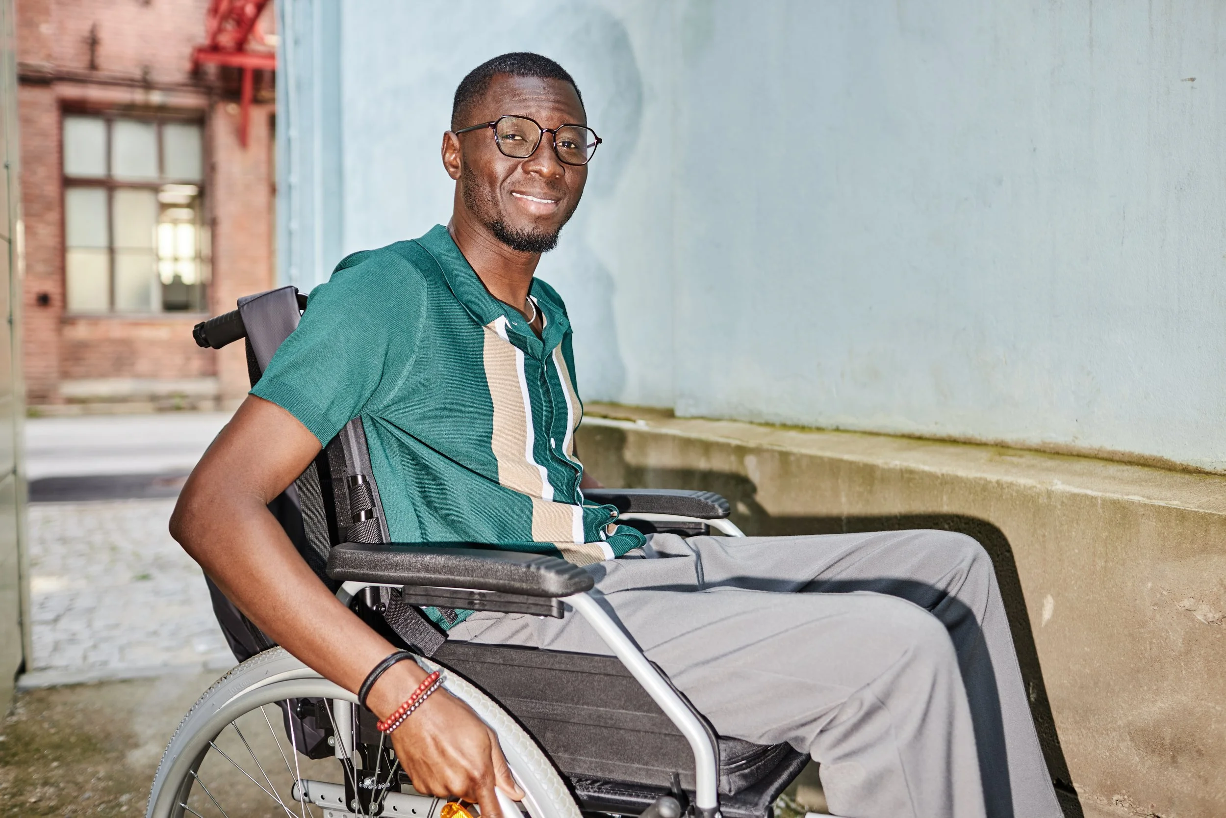 Person in a wheelchair smiling outdoors, wearing a green shirt with beige stripes and gray pants.