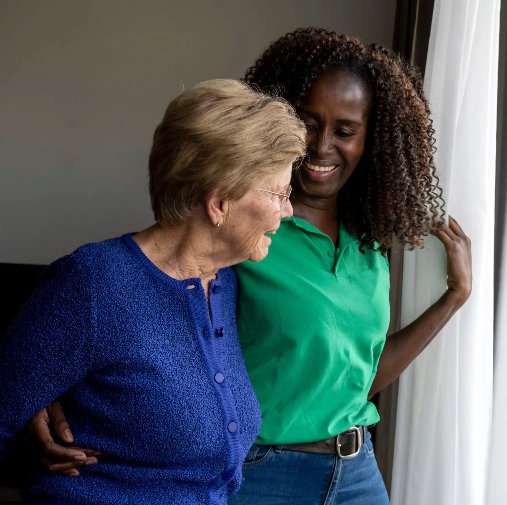 An older adult in a blue sweater and a younger person in a green shirt stand by a window, smiling warmly at each other, conveying care and companionship.