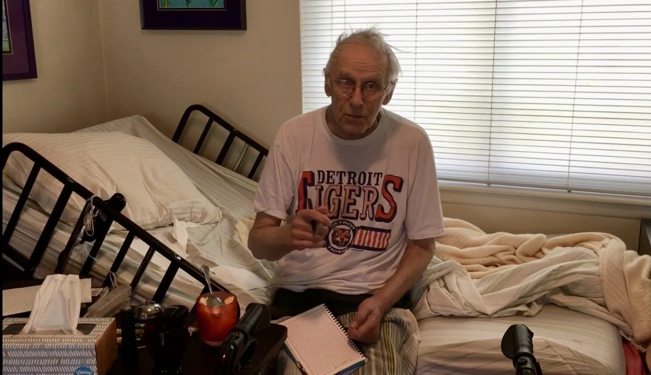 Man with white hair wearing glasses and a Detroit Tigers shirt sits on a hospital-type bed set up in a suburban dining room. He’s holding a pen with a notebook on his lap.