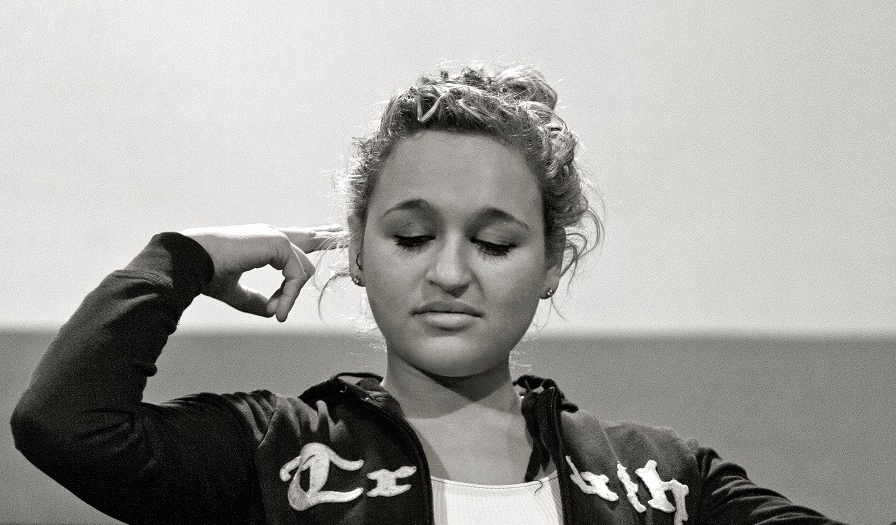 A young woman is captured using sign language with her eyes closed. She wears minimal eye makeup and has a few ear piercings. She wears a zip up hoodie and shirt underneath.