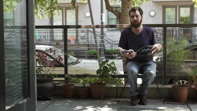 A man sits on a patio surrounded by plants. He has light skin, brown hair and a beard. He is wearing a dark t-shirt and jeans. He is holding a watering can.