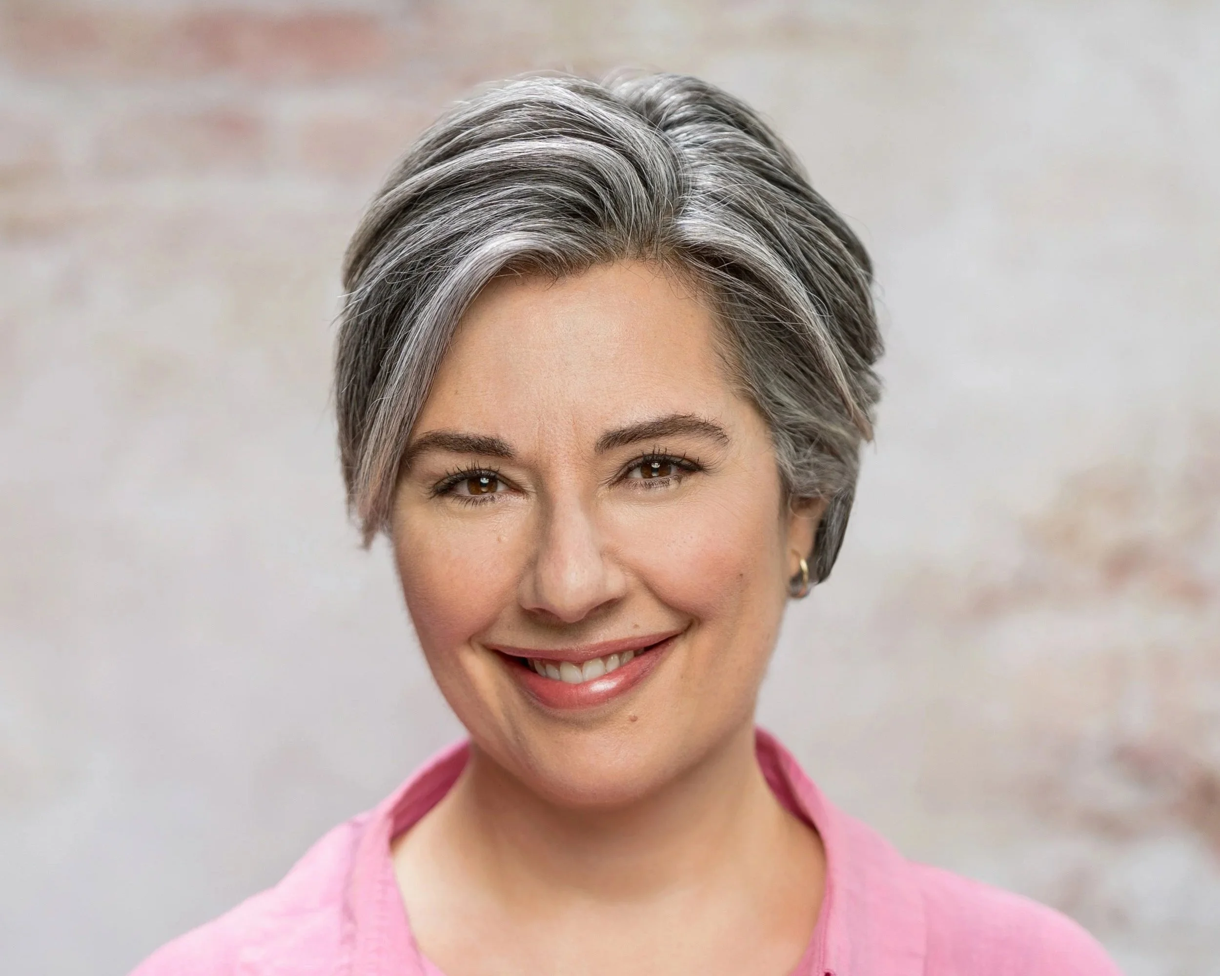 Close-up portrait of a smiling woman with short, gray and black hair, wearing a pink shirt, against a neutral background.