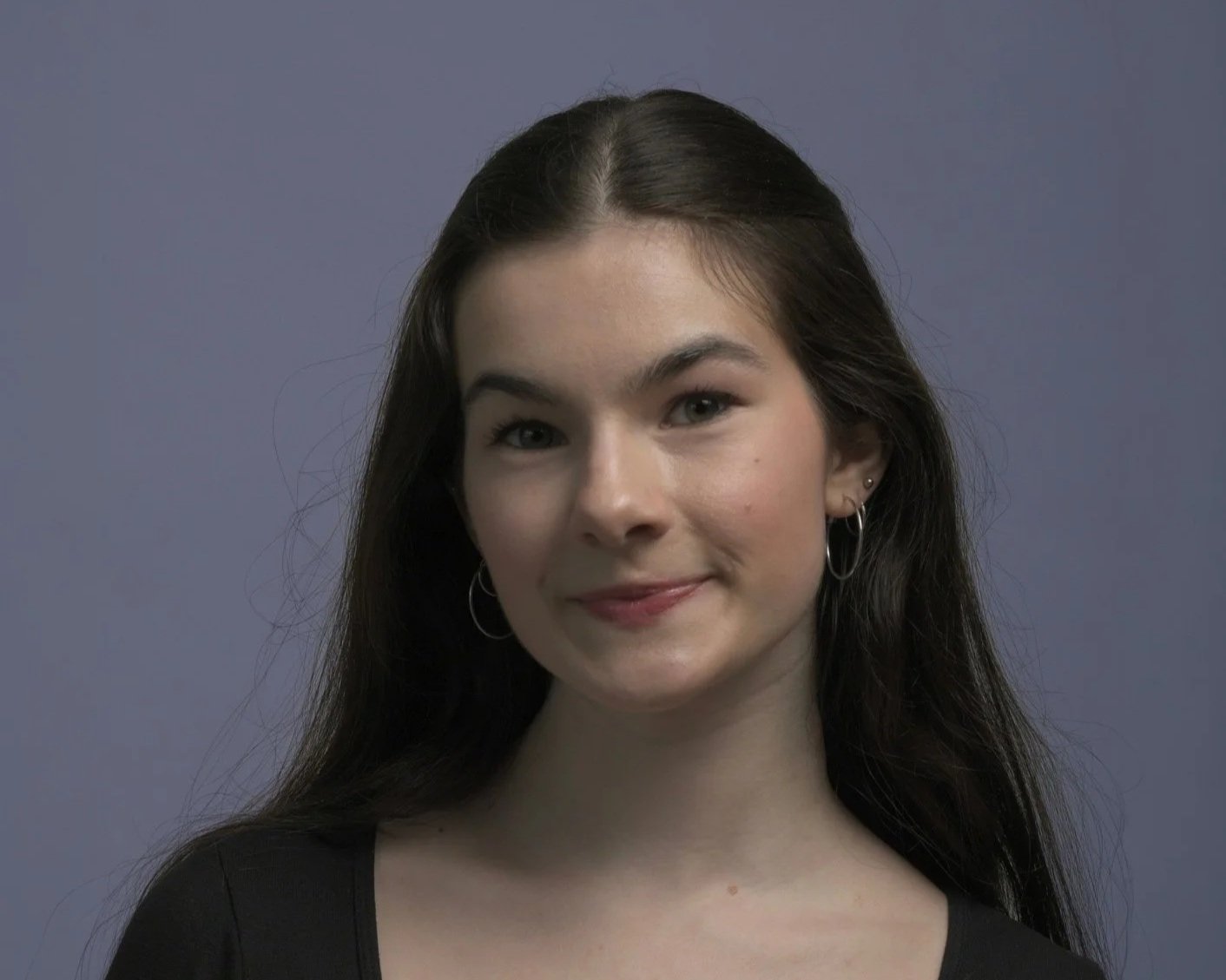 A young woman with long dark hair, wearing hoop earrings and a black top, smiling slightly at the camera, against a muted gray background.