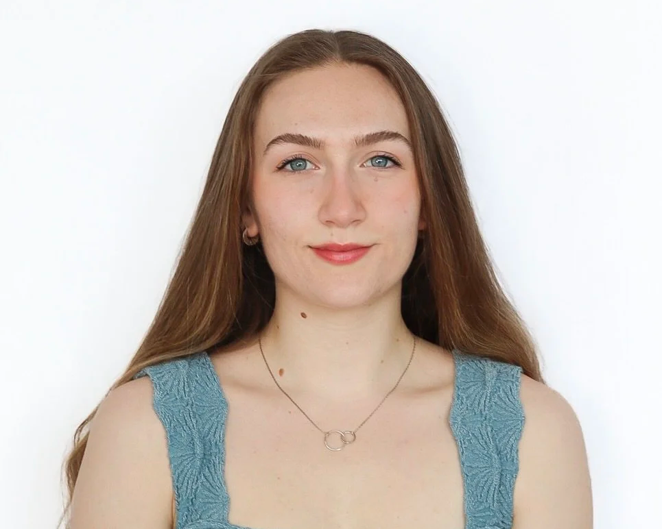 A young woman with long, straight, reddish-brown hair, wearing a sleeveless teal top with textured fabric and a silver necklace with two interlinked circles, smiling softly against a plain white background.