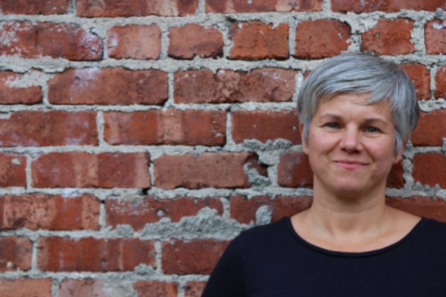 A woman with short gray hair smiling, standing in front of a red brick wall.