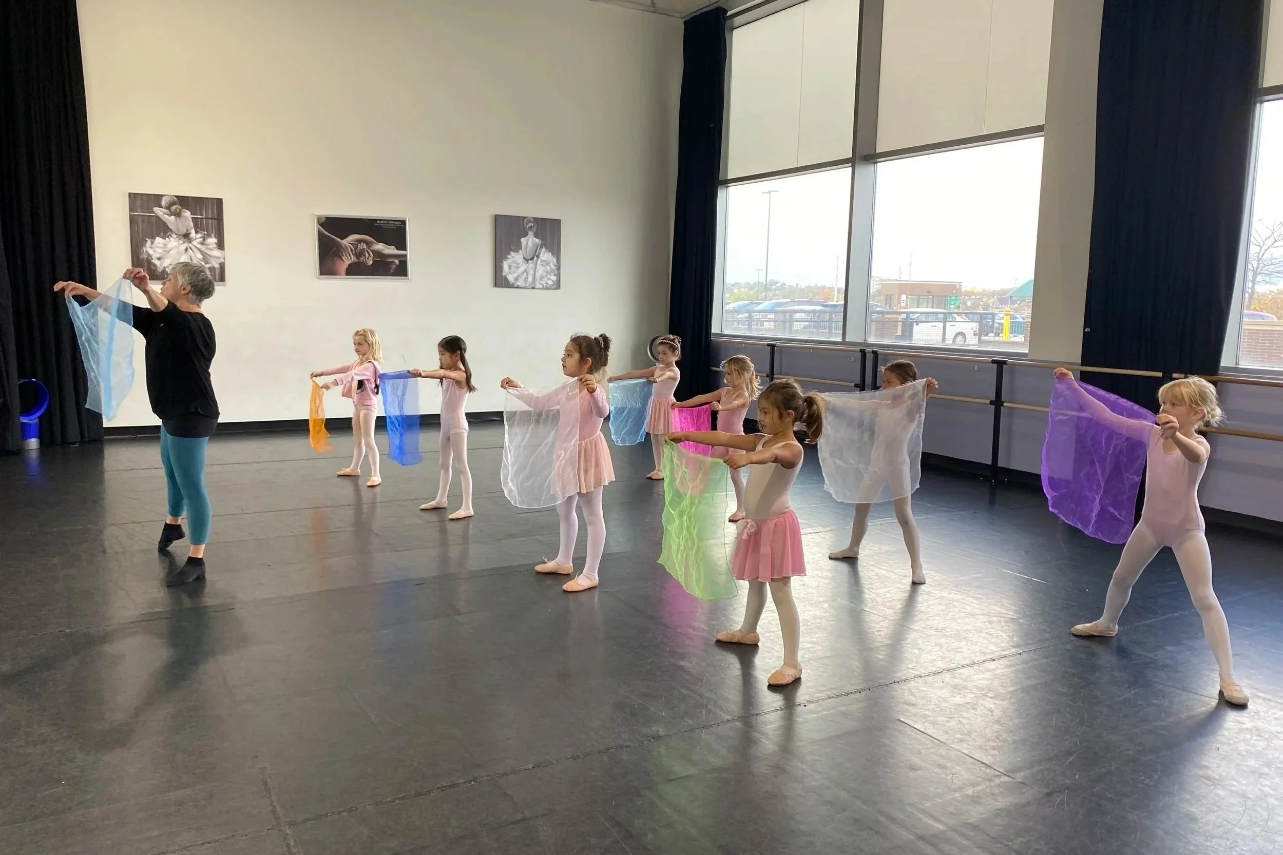 A ballet teacher instructs young girls in a ballet class, with each girl holding colorful scarves while standing in a line in a dance studio with large windows and ballet barres.