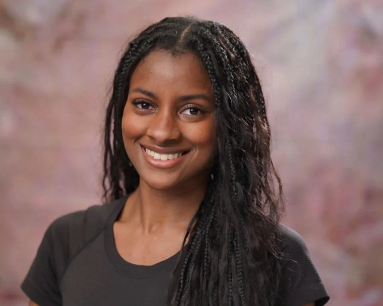 A woman with long braided hair smiling at the camera, wearing a black t-shirt, standing against a soft pink and purple background.