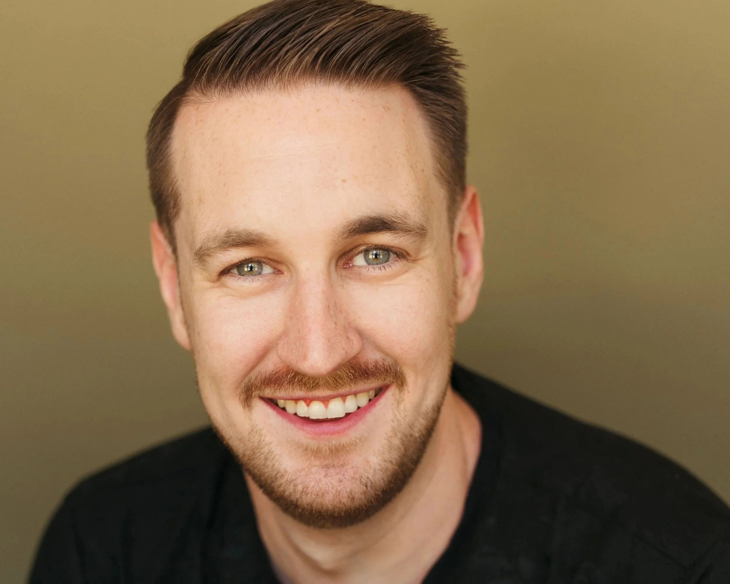 A young man with light skin, short brown hair, and blue eyes smiling at the camera, wearing a black shirt, against a muted green background.