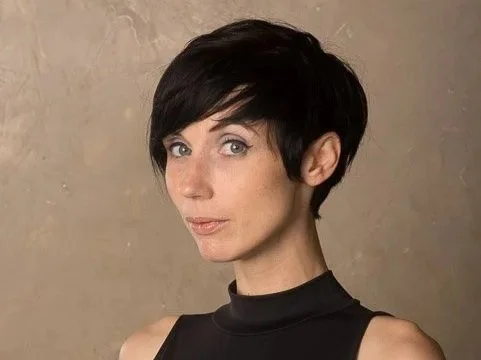 A woman with short black hair wearing a sleeveless black top poses against a beige background.