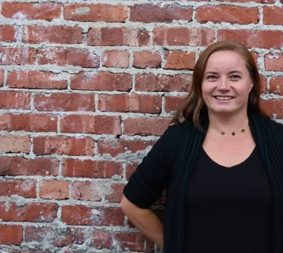 A woman with shoulder-length brown hair smiling, standing in front of a red brick wall, wearing a black top and a black cardigan.