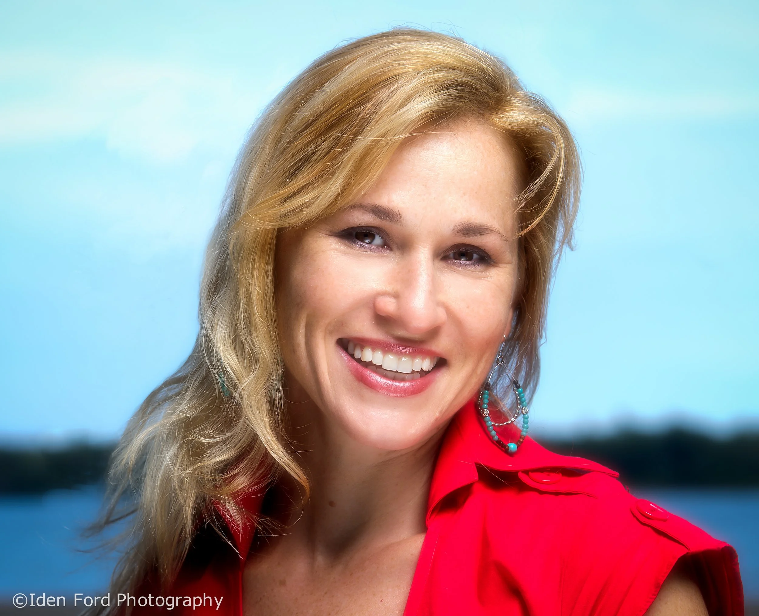 Close-up of a woman's face smiling, with blond hair, wearing a red blazer, turquoise necklace, and earrings, against a blue background.