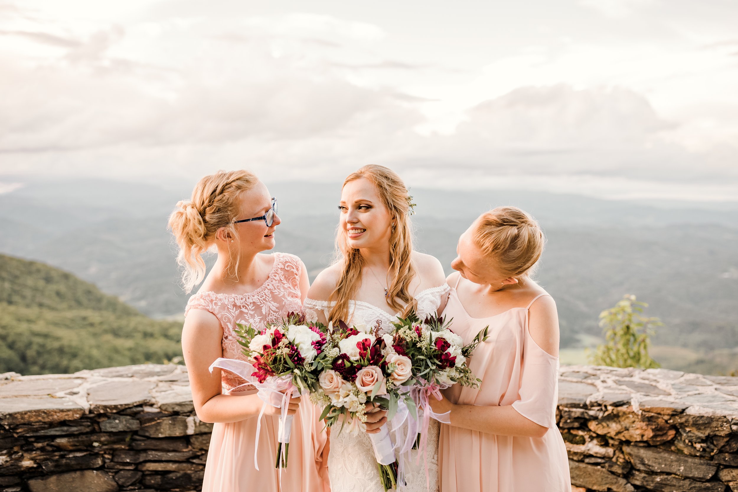 Bride with two bridesmaids holding bouquets in front of a mountain view stone wall at sunset