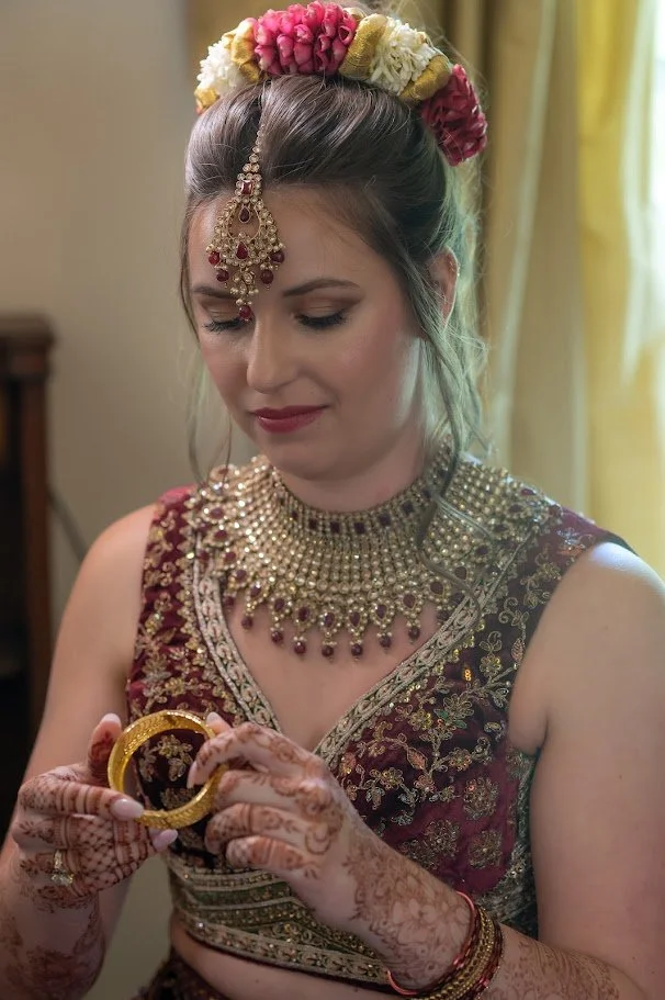 Close-up of a bride in ornate attire and jewelry looking down, holding a bangle, with floral hair adornments.