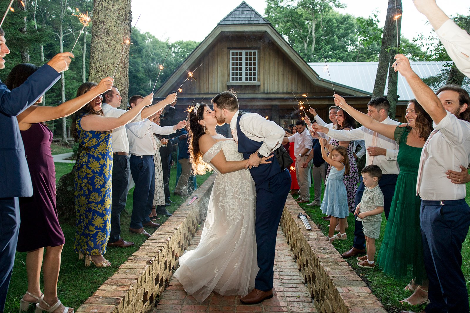Bride and groom kissing during a sparkler exit on a brick walkway surrounded by wedding guests.