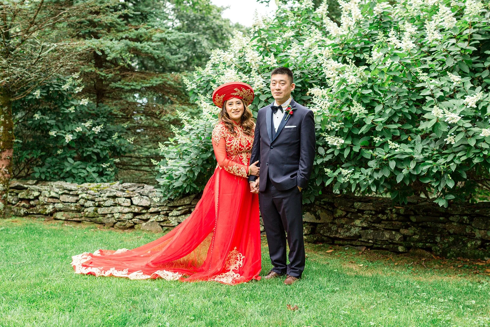 Bride in a red traditional wedding dress standing with groom in a black tuxedo for an outdoor garden portrait.