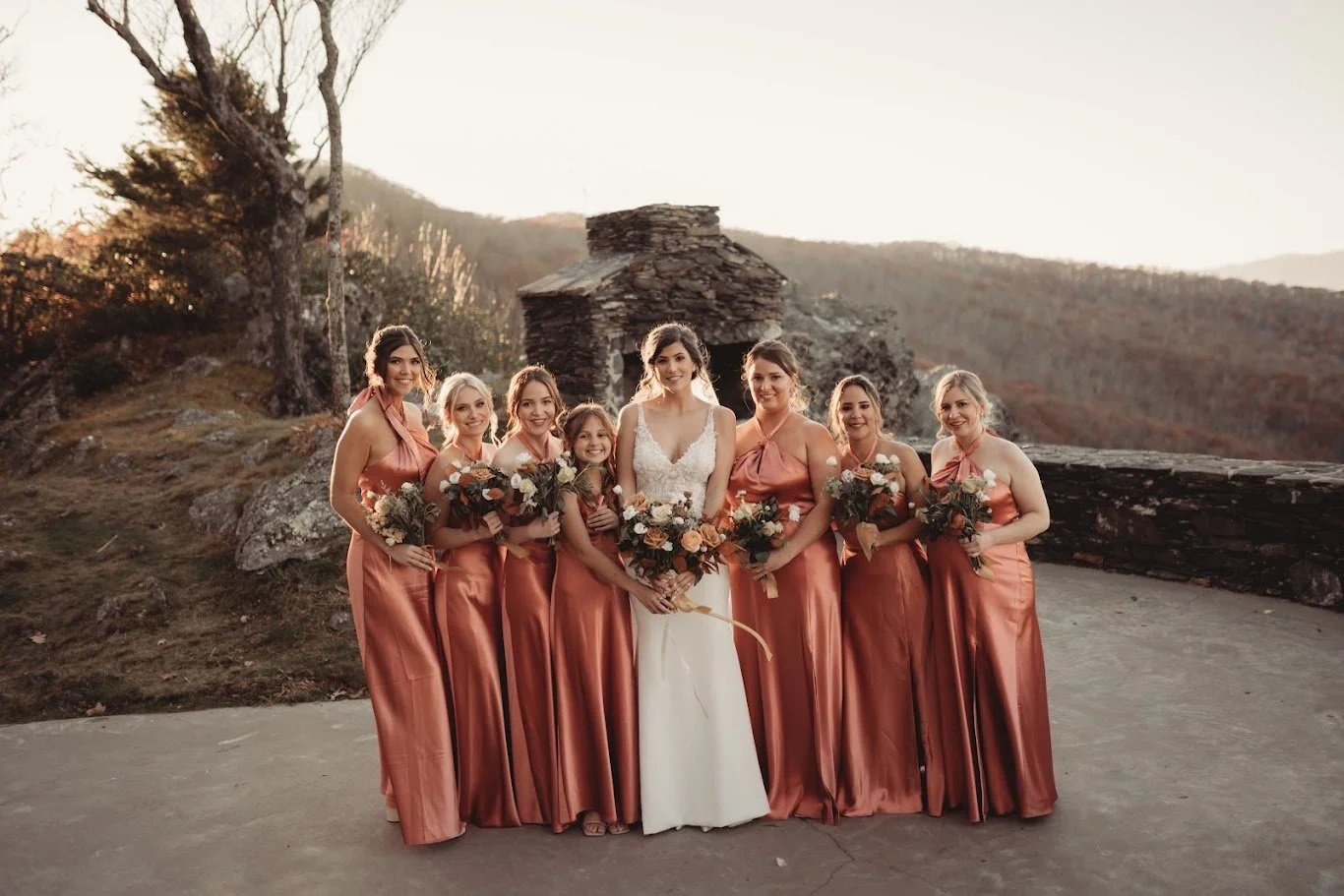 Bride posing with bridesmaids in satin dresses holding bouquets with a mountain overlook at sunset