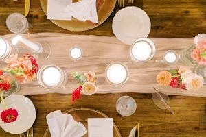An overhead view of an elegant wooden table set with a sheer peach runner, candles in glass holders, floral arrangements, gold chargers, and white plates.