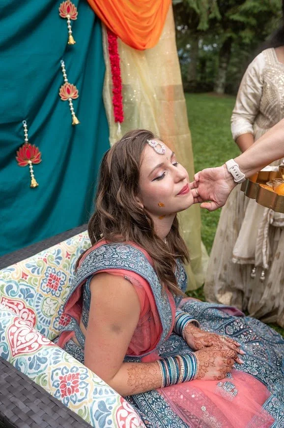 Person receives turmeric paste on their face during a pre-wedding ceremony while guests watch outdoors.