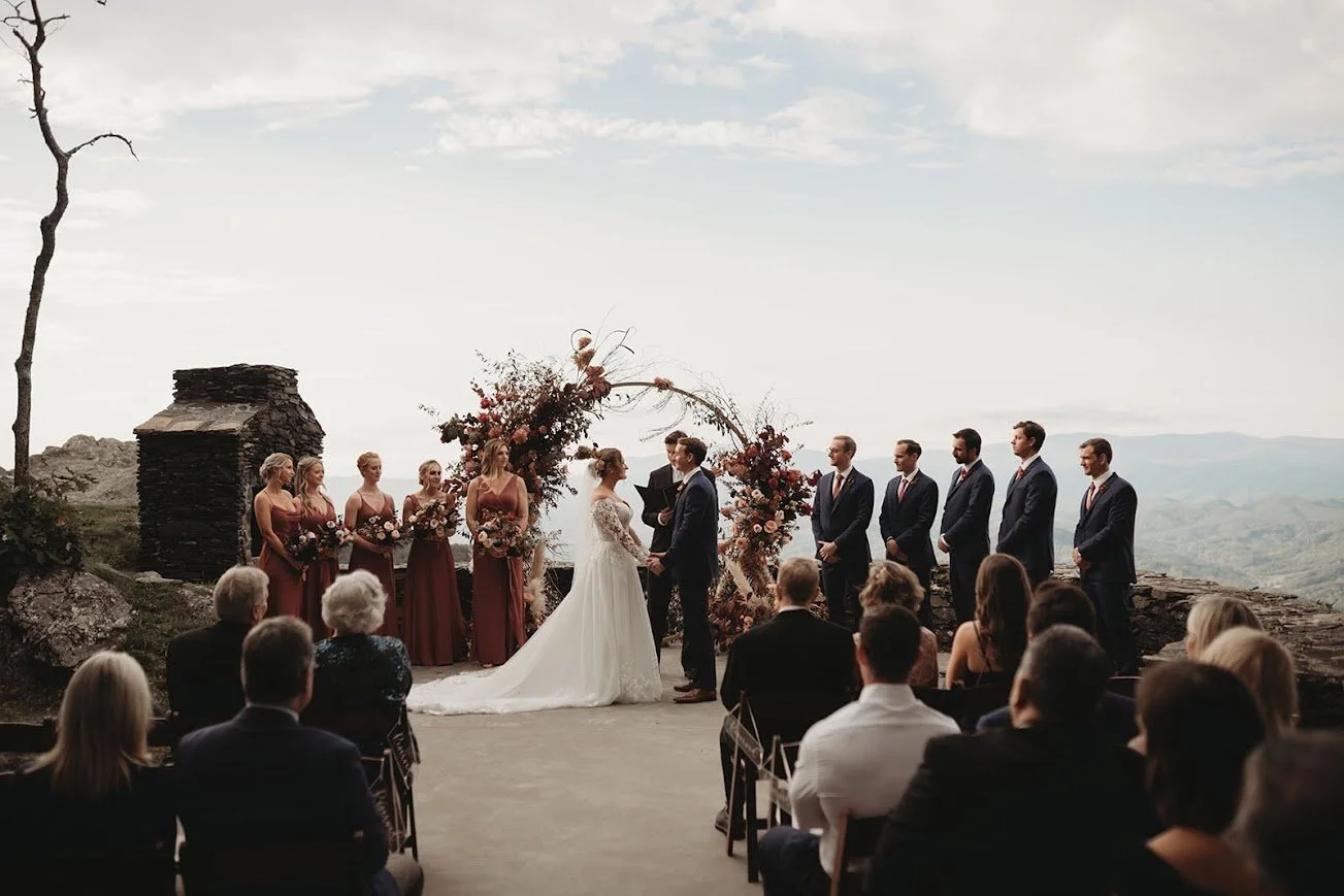 A scenic wedding ceremony at a mountain overlook with a circular floral arch and seated guests.