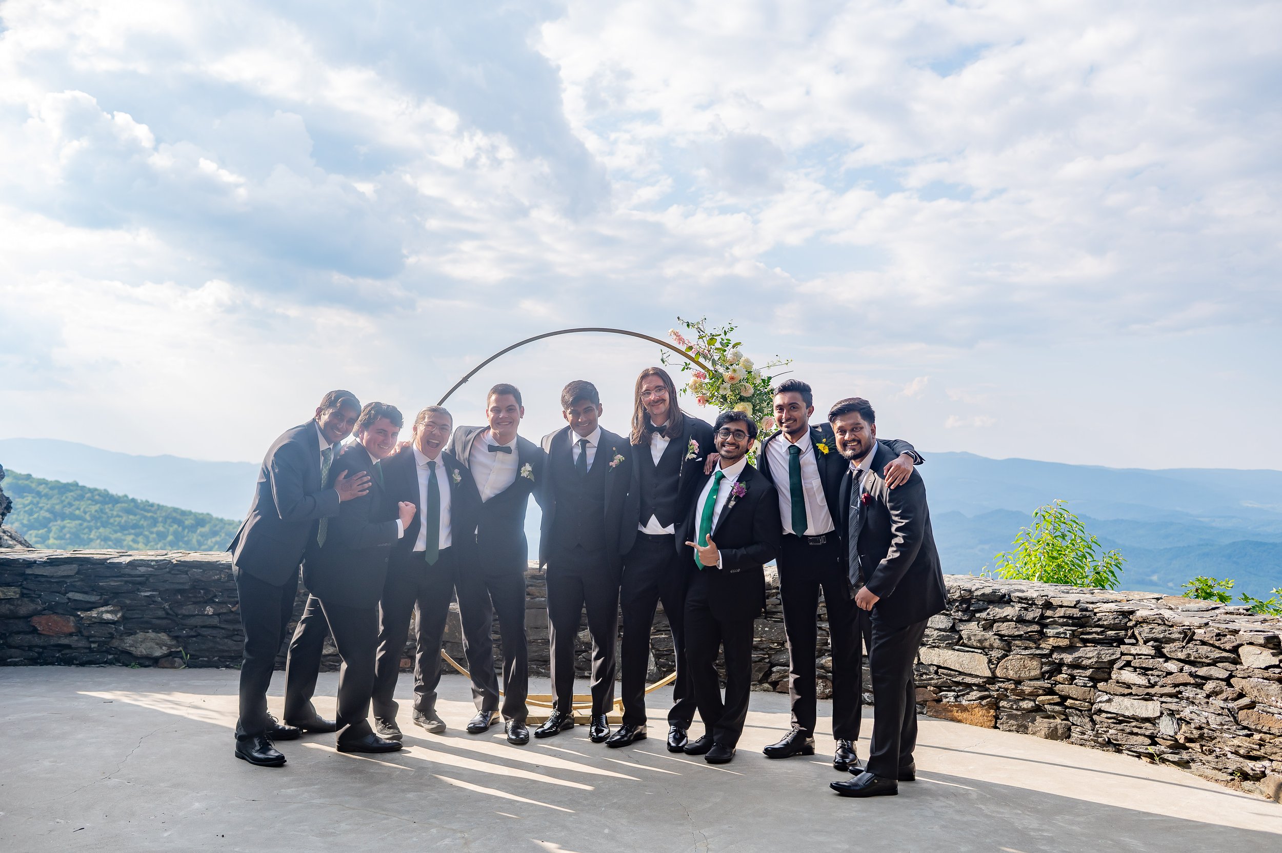 Groom and groomsmen in black suits posing together at a mountain overlook wedding venue.