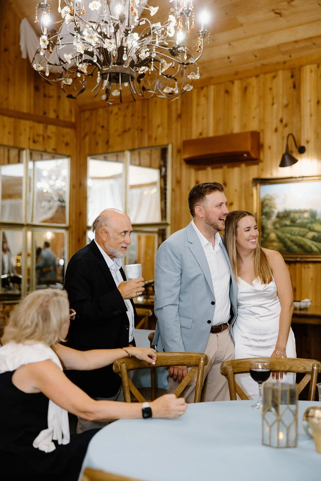 Wedding guests smiling and watching a toast in a rustic wood-paneled reception space under a decorative chandelier.