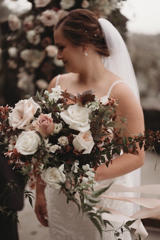 Bride in a wedding gown and veil holding a large bouquet of white and blush roses in soft natural light