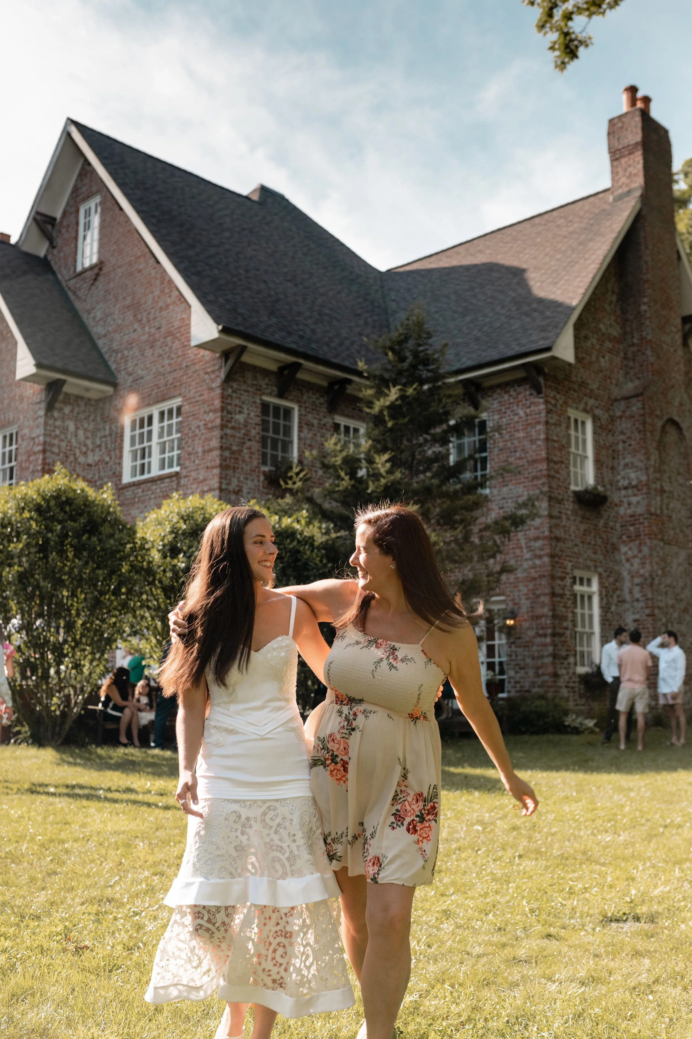 Two guests walking together on a sunny lawn outside a brick estate wedding venue during a weekend celebration.