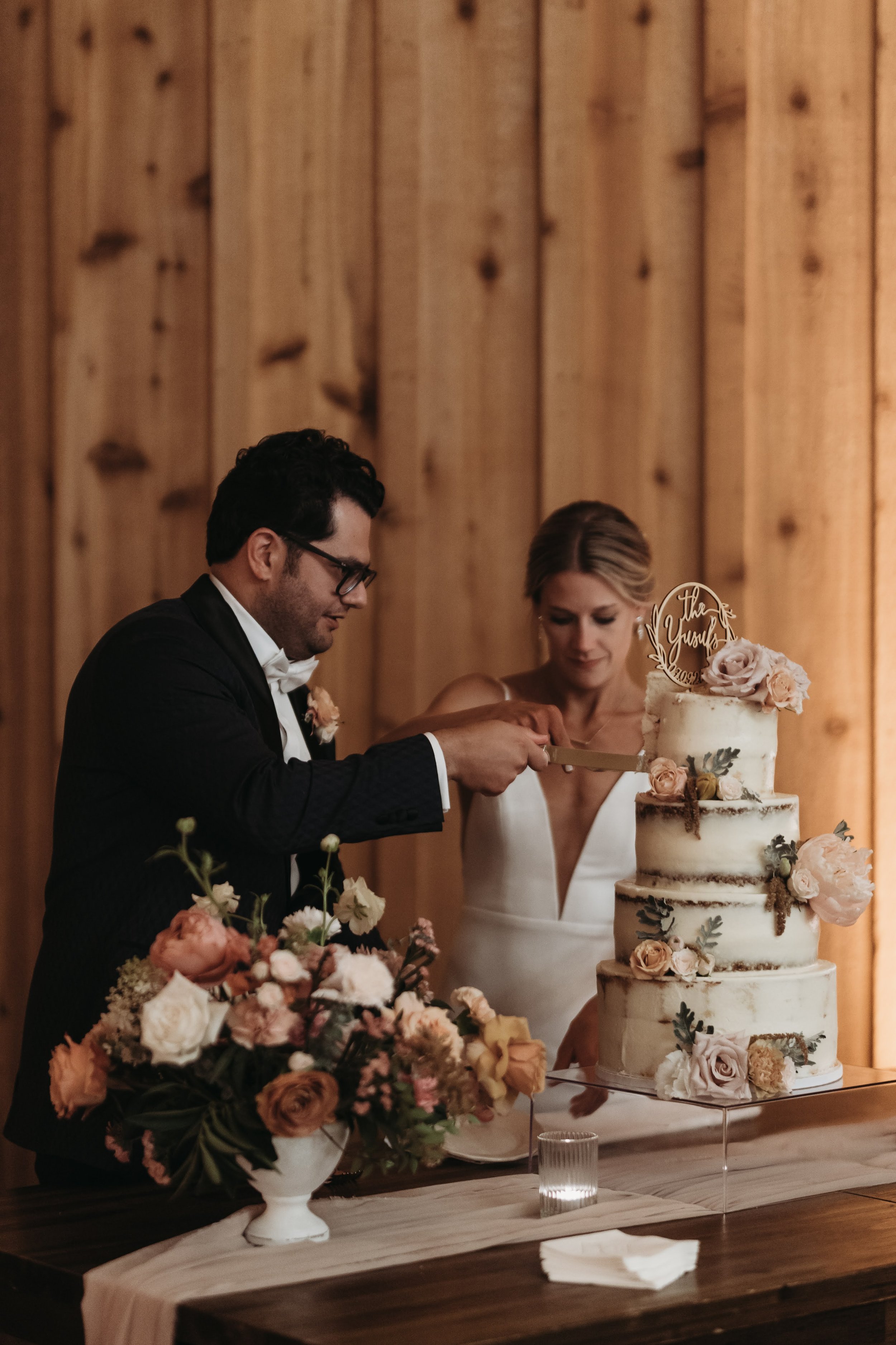 Bride and groom cutting a tiered floral wedding cake inside a warm wood reception venue.