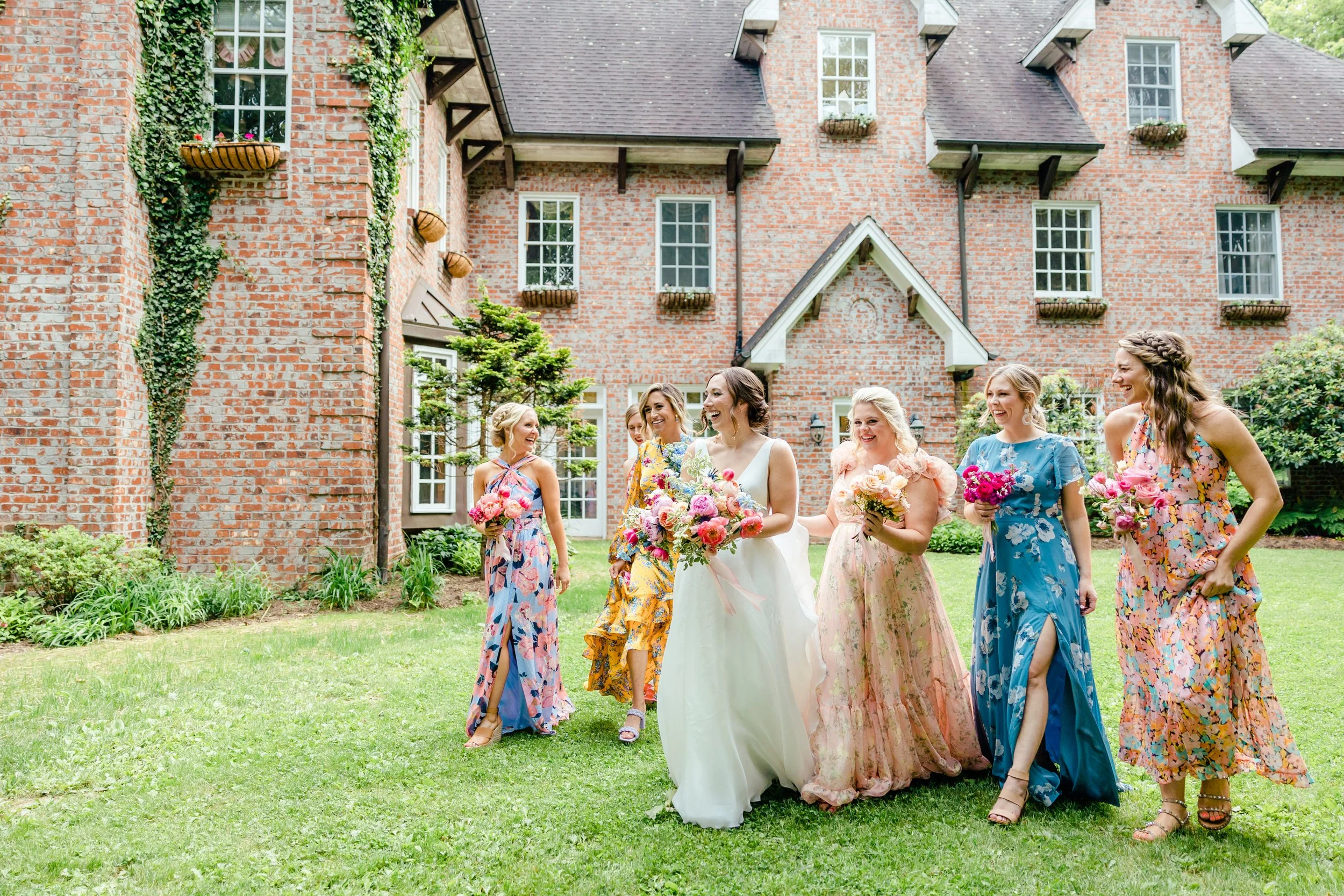Bride walking with bridesmaids in colorful floral dresses outside a historic brick estate wedding venue.