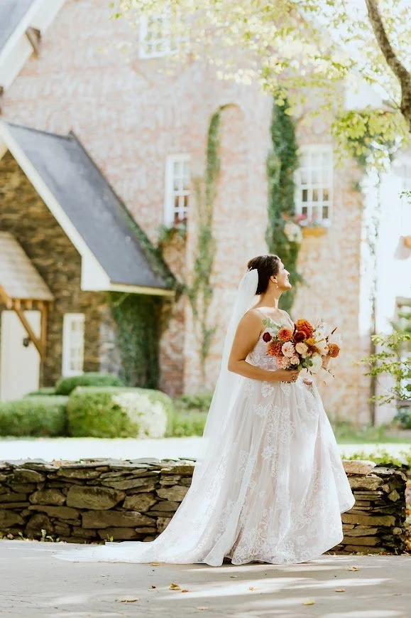 Bride in a lace gown holding a bouquet beside a low stone wall in the Twickenham House & Hall courtyard