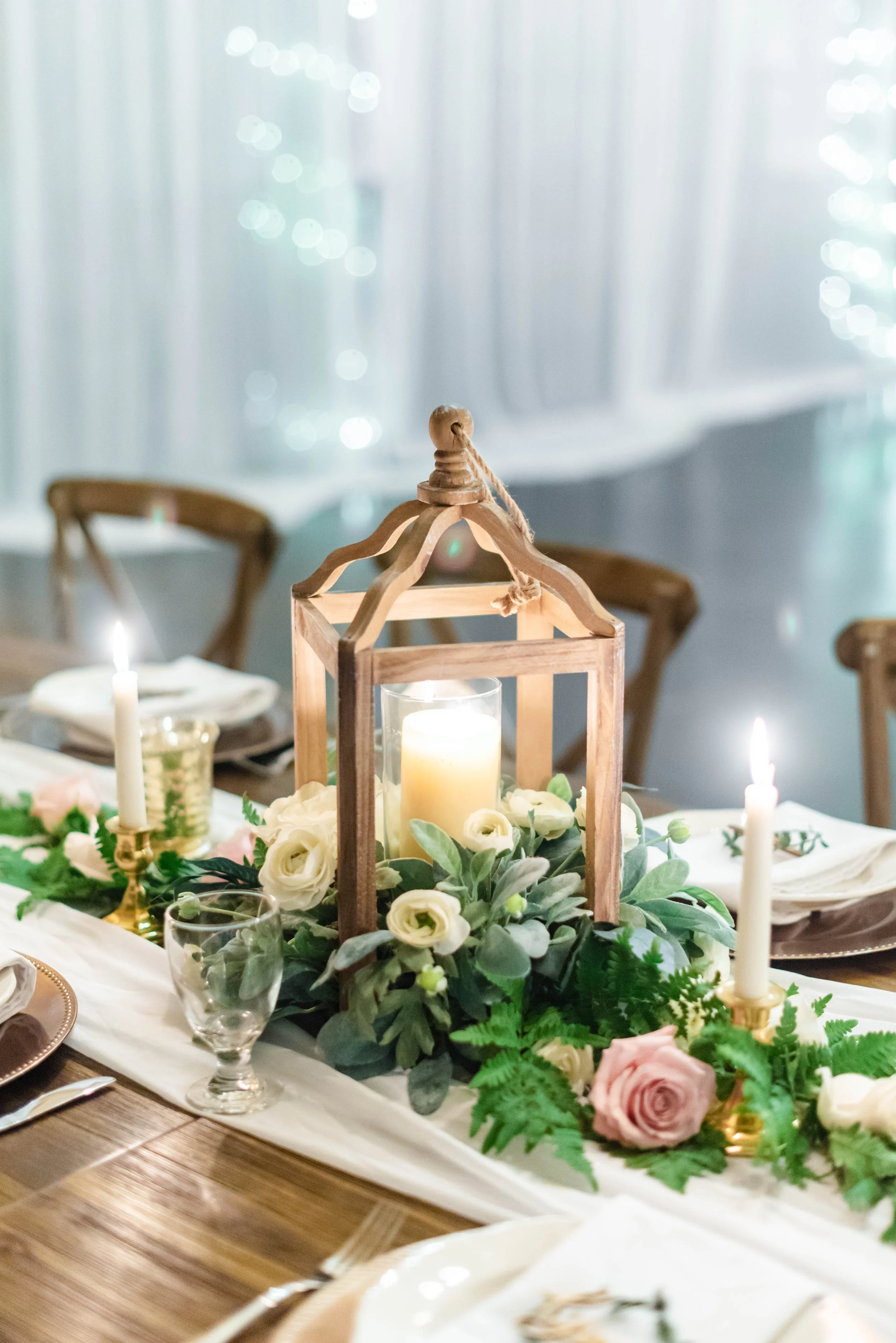 Wooden lantern centerpiece with a pillar candle surrounded by white blooms, blush rose, and lush greenery, flanked by taper candles on a reception table.