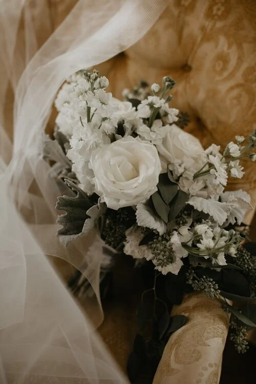 An elegant white wedding bouquet with roses, gypsophila, and greenery rests on a vintage gold chair, beside a wedding veil.